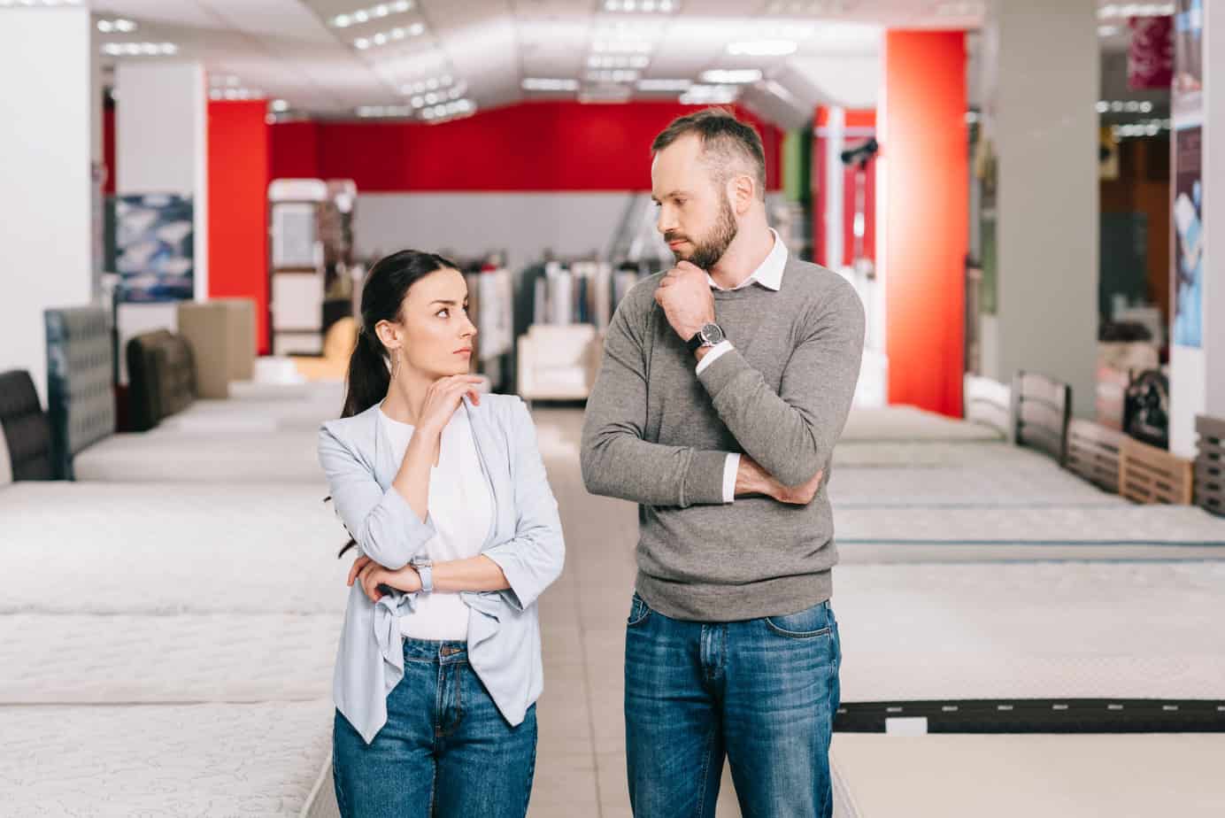 Pensive couple discussing mattress options in a furniture store, surrounded by various mattresses, reflecting the personalized shopping experience offered by ComfyQuiz.