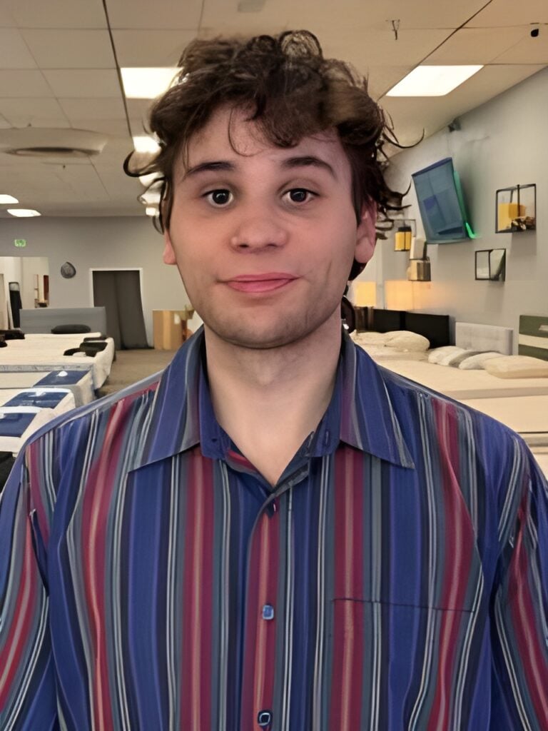 Young man in a striped shirt standing in a mattress showroom with beds displayed in the background, representing the team at Boise Mattress focused on enhancing sleep quality.