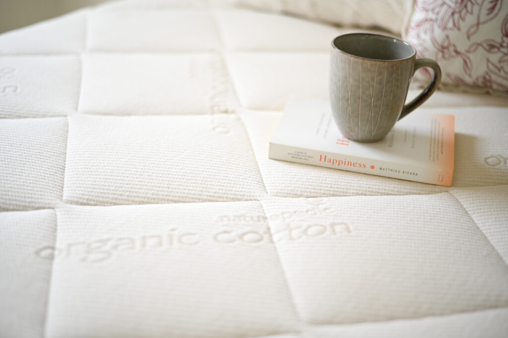 Organic cotton mattress by Naturepedic with a coffee mug and book titled "Happiness," emphasizing a cozy, non-toxic sleep environment.